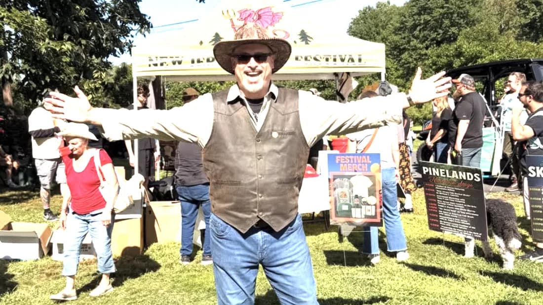 Professor B standing on grass with both arms outstretched at an outdoor festival, in front of a white event tent and vendor signage.